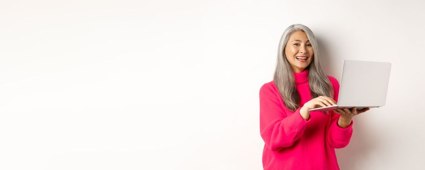 Beautiful asian senior woman entrepreneur working with laptop, laughing and smiling at camera, standing over white background.