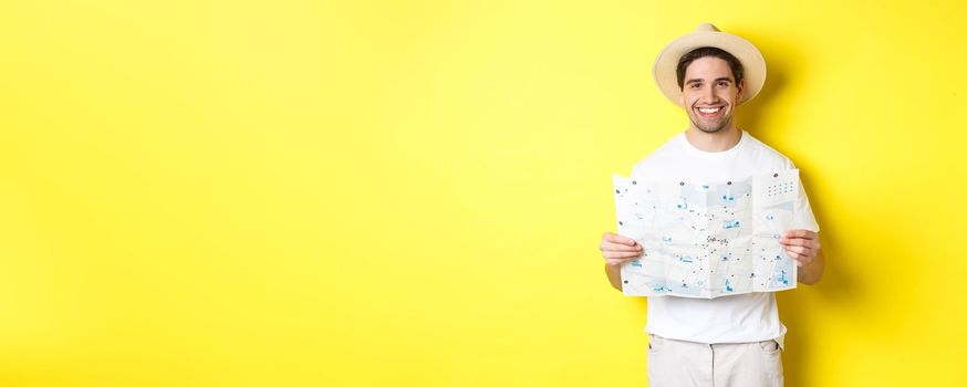 Travelling, vacation and tourism concept. Smiling young man going on trip, holding road map and smiling, standing over yellow background.