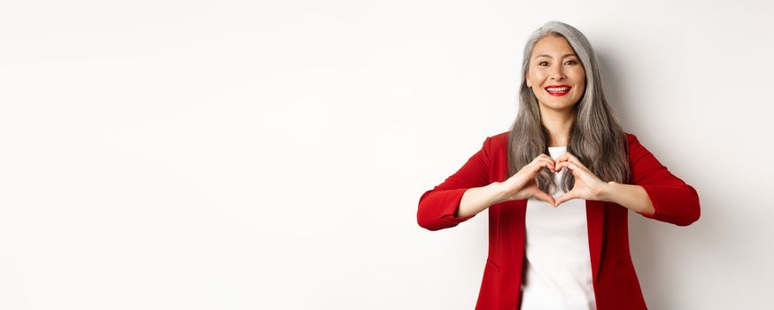 Beautiful asian mature woman in red blazer and makeup, showing heart sign and smiling, I love you gesture, standing over white background.