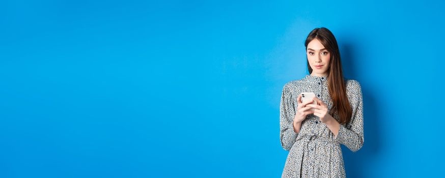 Stylish modern woman in dress, chatting on smartphone and smiling at camera, standing on blue background.