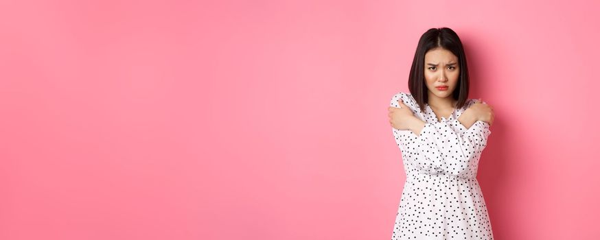 Timid and offended asian girl cross arms on chest, staring defensive and insulted at camera, standing in dress over pink background.