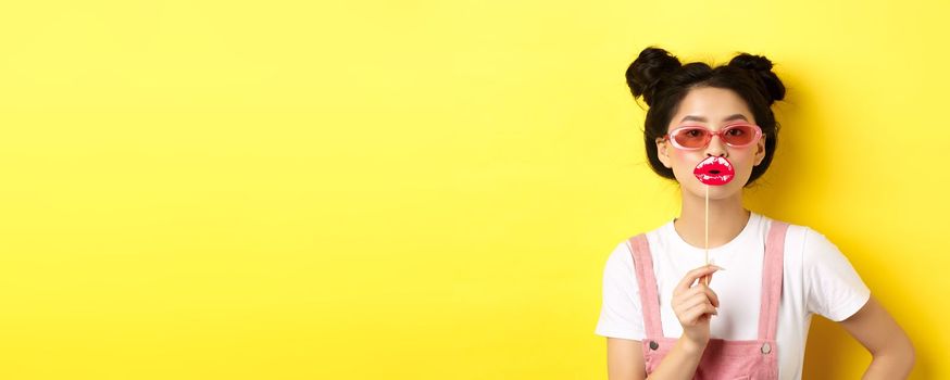 Summer and fashion concept. Stylish asian glam girl in sunglasses, holding party lips mask, standing on yellow background.