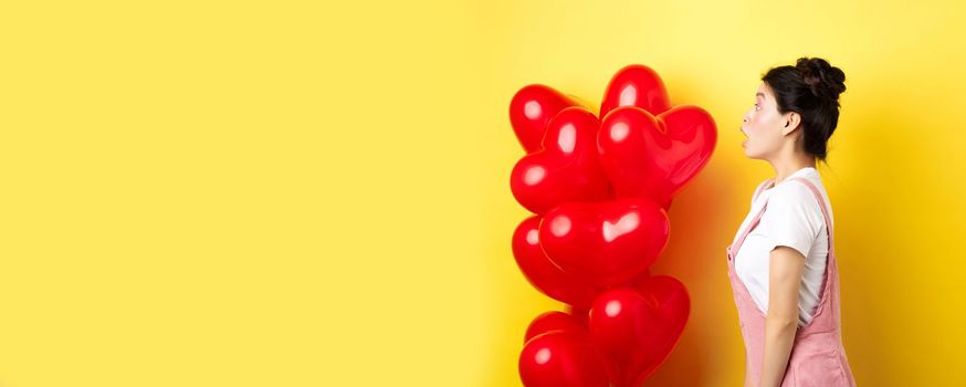 Valentines day and relationship concept. Profile of surprised asian girl staring left at logo, standing near heart balloons, yellow background.