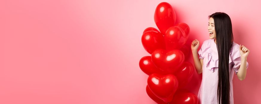 Asian teenage girl with long hair, cheering from valentines day romantic gift, looking at logo and smiling happy, jumping from joy near lovers gift heart balloons, pink background.