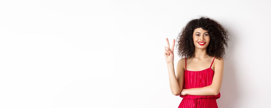 Attractive female model in red dress and makeup, showing number two and smiling, standing over white background.