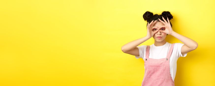 Funny asian girl look through hand binoculars, watching you, standing in summer clothes on yellow background.