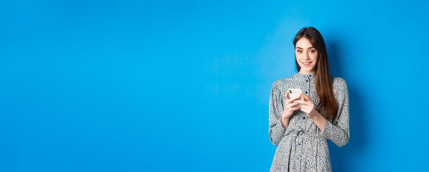 Attractive caucasian woman in dress using mobile phone and smiling, standing in dress against blue background.