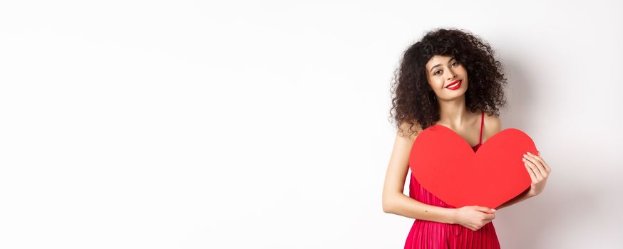 Romantic tender woman with curly hair, hugging big red heart and smiling, look with love, standing against white background.