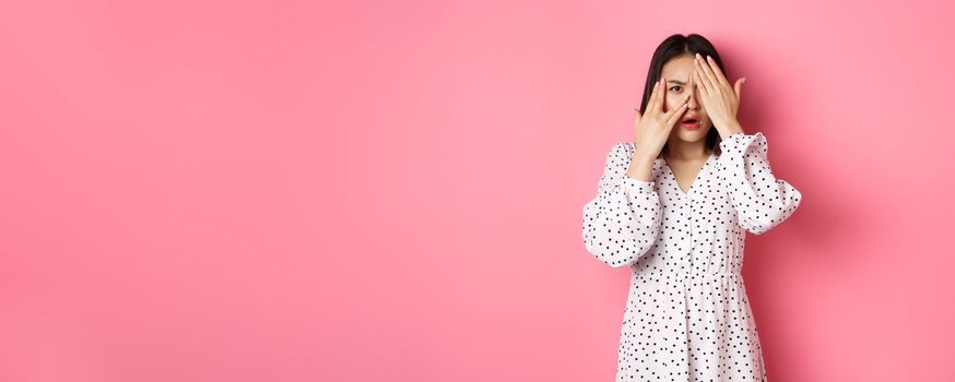 Disappointed asian woman peeking through fingers, open eyes and frowning displeased, staring with disdain at camera, standing over pink background.