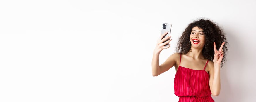 Elegant young woman in red dress taking selfie on smartphone, posing on event party, standing with mobile phone on white background.