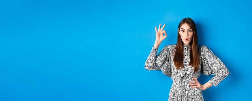 Impressed caucasian woman in dress showing okay sign, look with amazement and say wow, praise awesome product, standing on blue background.