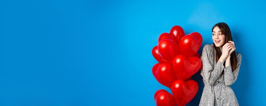 Valentines day. Beautiful romantic girl dreaming off date, standing near lovely heart balloons and smiling, blue background.