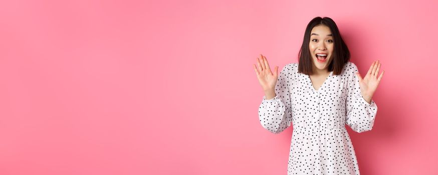 Happy and surprised asian girl receive surprise, looking glad hearing good news, standing amazed in dress against pink background.