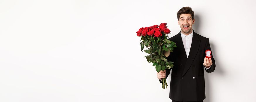 Handsome smiling man in black suit, holding roses and engagement ring, making a proposal to marry him, standing against white background.