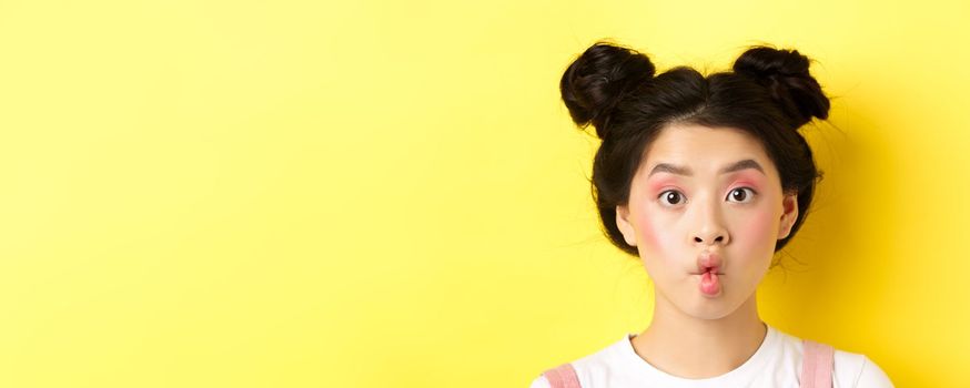Close-up of teen korean girl pucker lips and looking funny at camera, standing with glamour makeup and stylish hairstyle, yellow background.