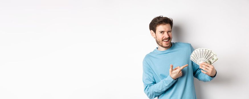 Handsome rich guy winking at camera, showing income, pointing finger at earned money and smiling, standing over white background.