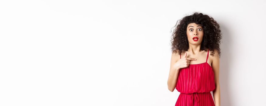 Surprised young woman in red dress pointing at herself, being confused and shocked, standing on white background. Copy space