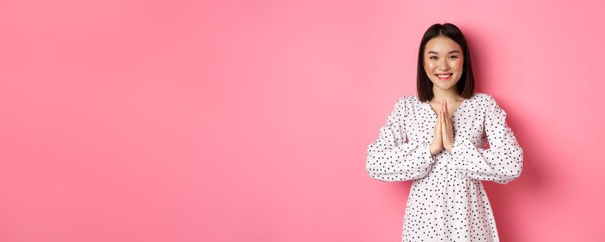 Beautiful asian woman thanking you, holding hands together in appreciation gesture, smiling happy at camera, standing grateful over pink background.