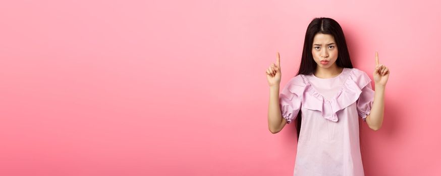 Sad and moody asian girl look from under forehead and sulking, pointing fingers up and complaining, staring jealous or upset, standing against pink background.