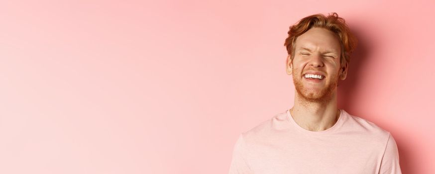 Close up of carefree man with red hair and beard, laughing and feeling happy, standing over pink background, having fun.