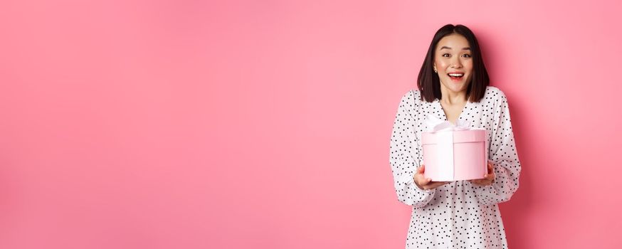 Surprised and happy asian woman receiving cute box with gift, standing amazed over pink background.