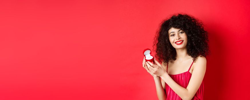 Romantic woman with curly hair, wearing red dress and showing engagement ring, standing happy on studio background.