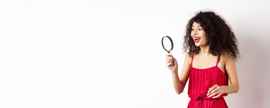 Excited woman in red dress look through magnifying glass and smiling, found interesting promo, standing on white background.