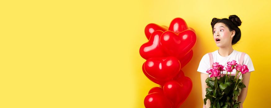 Happy Valentines day. Surprised asian girl looking left with silly face, holding cute pink flowers, receive romantic bouquet, standing on yellow background near red hearts balloons.