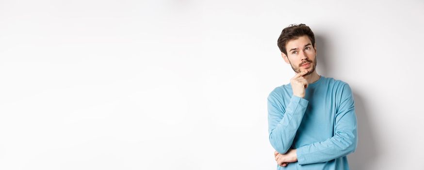 Image of handsome young man making choice, thinking and looking pensive up, standing over white background.