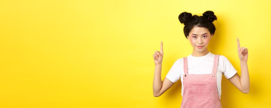 Confident asian young girl with beauty makeup, pointing fingers up, showing advertisement, standing on yellow background and smiling.
