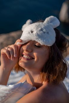 Young woman in sleep mask posing on the seashore. She is wrapped in a white blanket. The coast of the sea at a height, around the sea and rocks, dawn