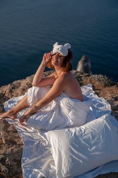 Young woman in sleep mask posing on the seashore. She is wrapped in a white blanket. The coast of the sea at a height, around the sea and rocks, dawn