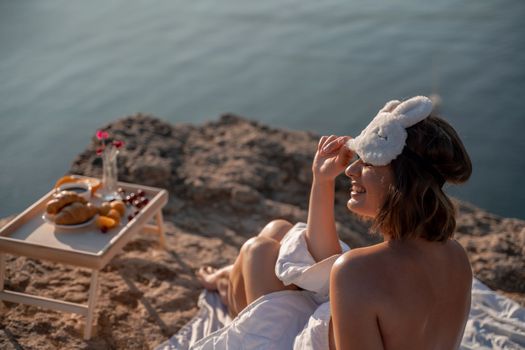 Young woman in sleep mask posing on the seashore. She is wrapped in a white blanket. The coast of the sea at a height, around the sea and rocks, dawn