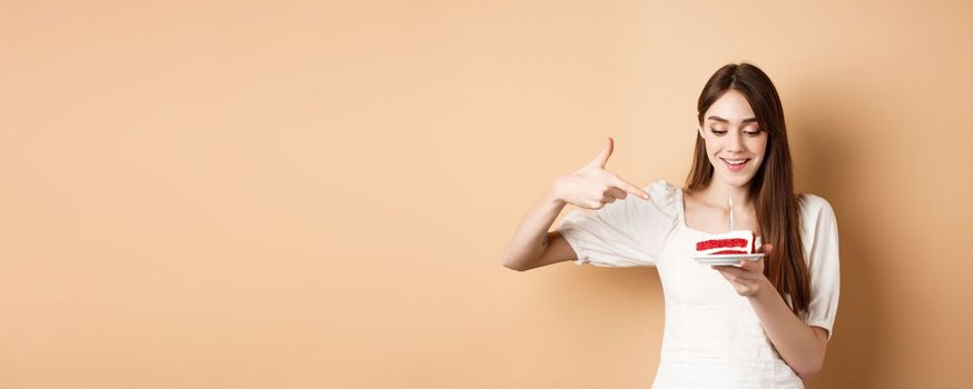 Cute girl pointing at birthday cake with candle and smiling excited, making bday wish, standing on beige background.