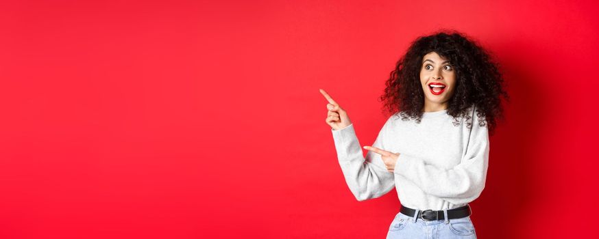 Excited pretty girl with curly hair and red lips, looking and pointing fingers left with amazed face, showing banner, standing on studio background.