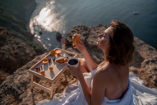 Woman covered with a blanket of bed relaxing and watching the seascape at sunrise. She holds a cup of coffee in her hand in front of her is a table with fruits and croissants. Wanderlust and freedom concept scene