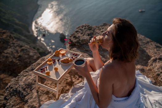 Woman covered with a blanket of bed relaxing and watching the seascape at sunrise. She holds a cup of coffee in her hand in front of her is a table with fruits and croissants. Wanderlust and freedom concept scene