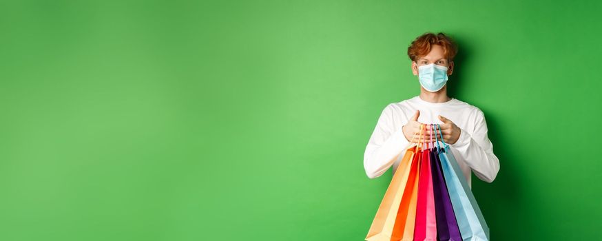 Pandemic and lifestyle concept. Cheerful redhead man going shopping in store, wearing medical mask and holding bags, standing over green background.