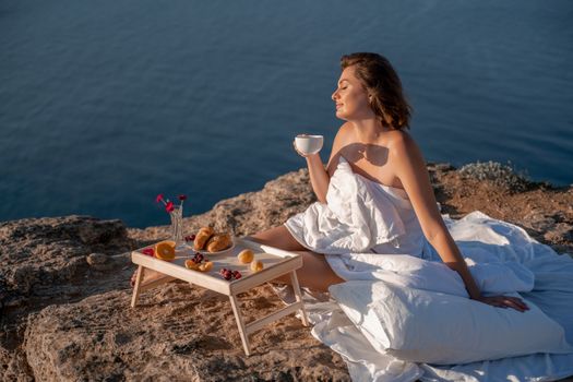 Woman covered with a blanket of bed relaxing and watching the seascape at sunrise. She holds a cup of coffee in her hand in front of her is a table with fruits and croissants. Wanderlust and freedom concept scene