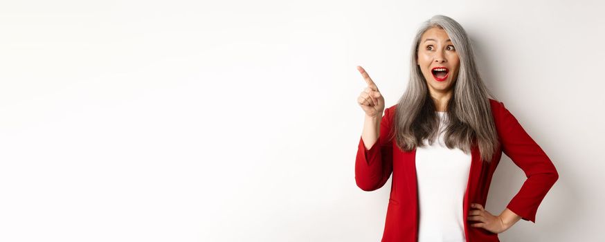 Surprised senior asian lady pointing upper left corner, checking out promo offer and smiling amazed, standing in red blazer over white background.