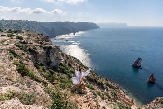 A beautiful young woman in a white light dress with long legs stands on the edge of a cliff above the sea waving a white long dress, against the background of the blue sky and the sea