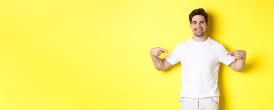 Happy attractive guy pointing fingers at your logo, showing promo on his t-shirt, standing over yellow background.