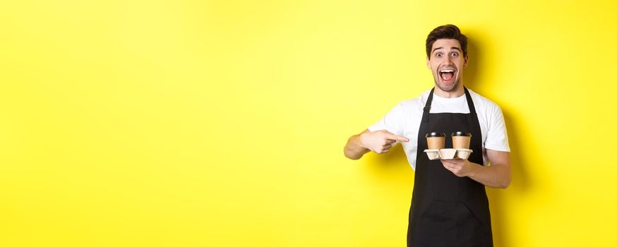 Excited barista in black apron pointing fingers at takeaway coffee cups, standing against yellow background happy.