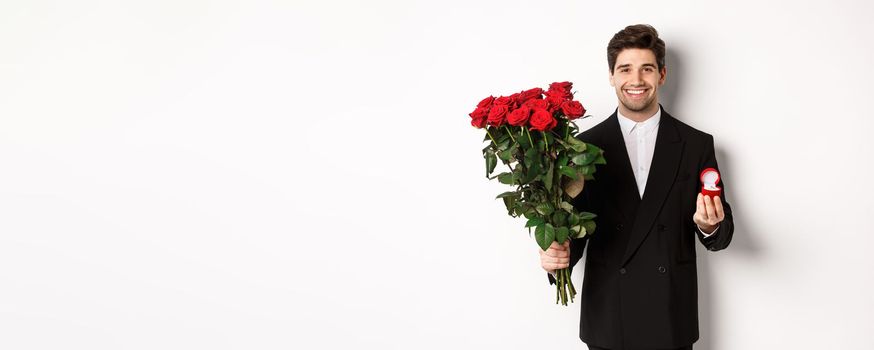 Handsome smiling man in black suit, holding roses and engagement ring, making a proposal to marry him, standing against white background.