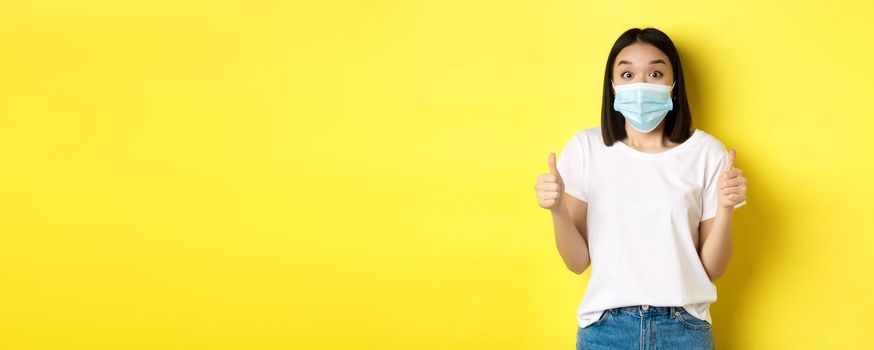 Covid-19, quarantine and social distancing concept. Cheerful asian woman in medical mask and white t-shirt showing thumbs up in approval, praise good deal, yellow background.