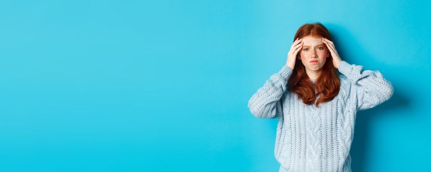 Distressed teenage redhead girl touching head, looking with troubled face expression, standing against blue background, have problem.