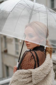 The blonde stands under a transparent umbrella during the rain. The fall season. Rear view. The woman is dressed in a black lace dress, her hair pulled back in a ponytail