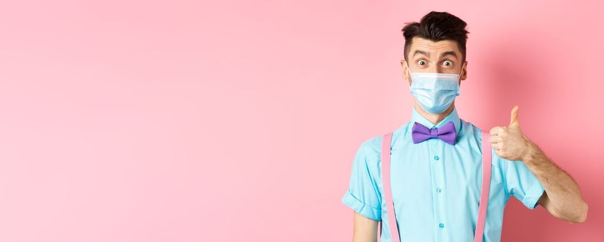 Coronavirus, healthcare and quarantine concept. Impressed young man in medical mask showing thumb up, looking satisfied, recommending good product, pink background.