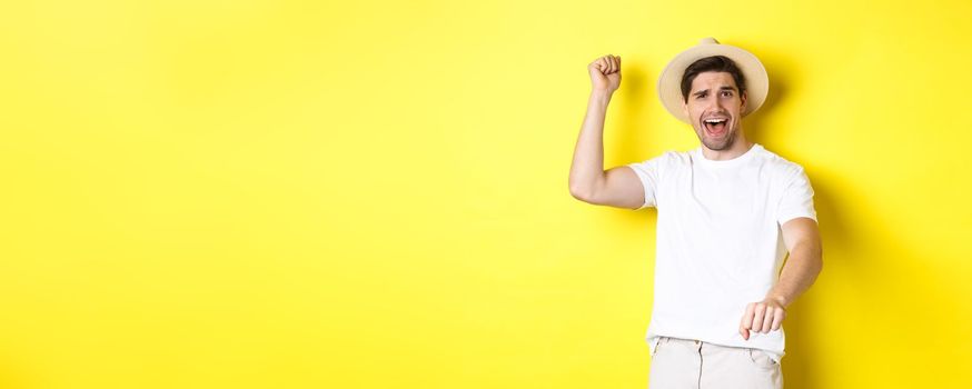 Concept of tourism and summer. Young man traveller showing rodeo gesture, standing in straw hat and white clothes, standing over yellow background.