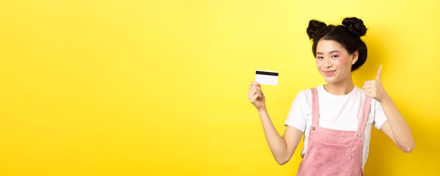 Shopping. Stylish asian glam girl showing thumb up and plastic credit card, standing on yellow background.